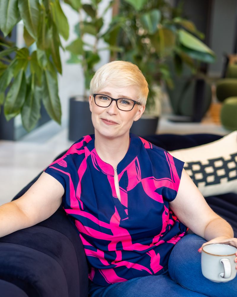Jenny Lucas, a woman with short blonde hair and dark-framed glasses, sitting on a navy sofa holding a mug and smiling at the camera