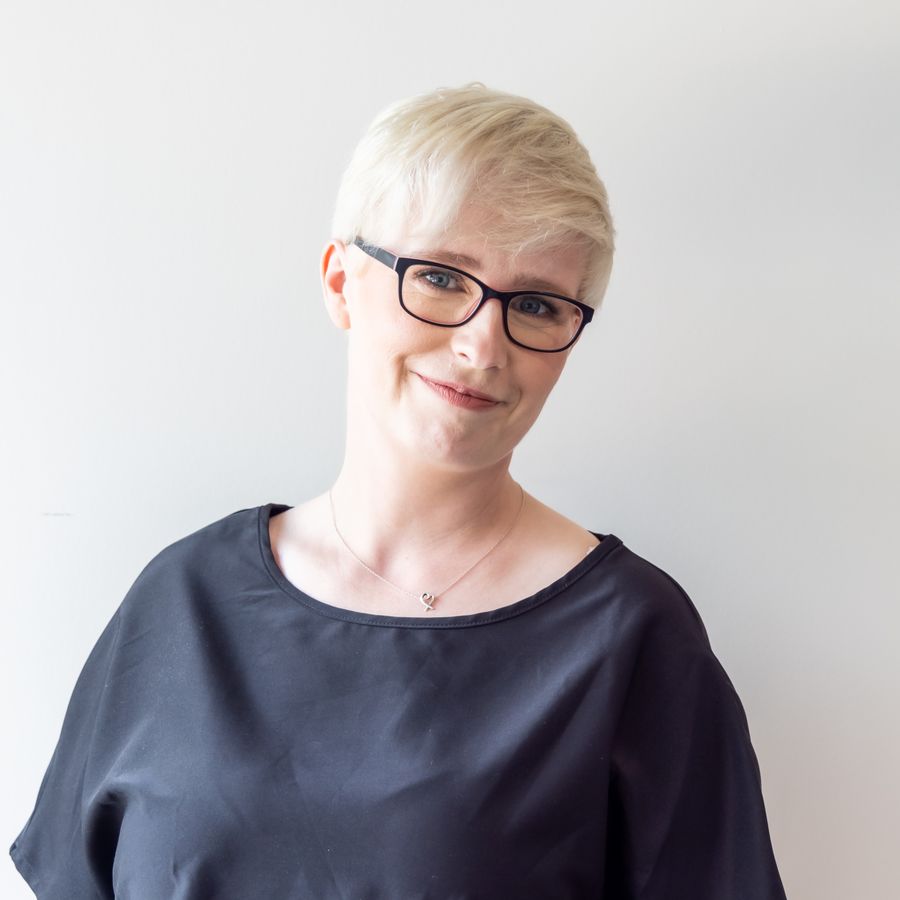 Jenny Lucas, a woman with short blonde hair and dark-framed glasses, wearing a black top against a plain pale background